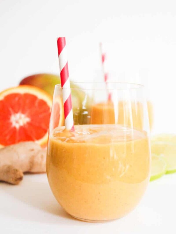Glass of orange smoothie with red and white spiral straw behind are ginger root , a mango, and a halved orange.
