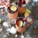 Overhead shot of three cranberry moscow mules in copper mugs with sugared cranberry swizzle sticks and bowl of sugared cranberries.