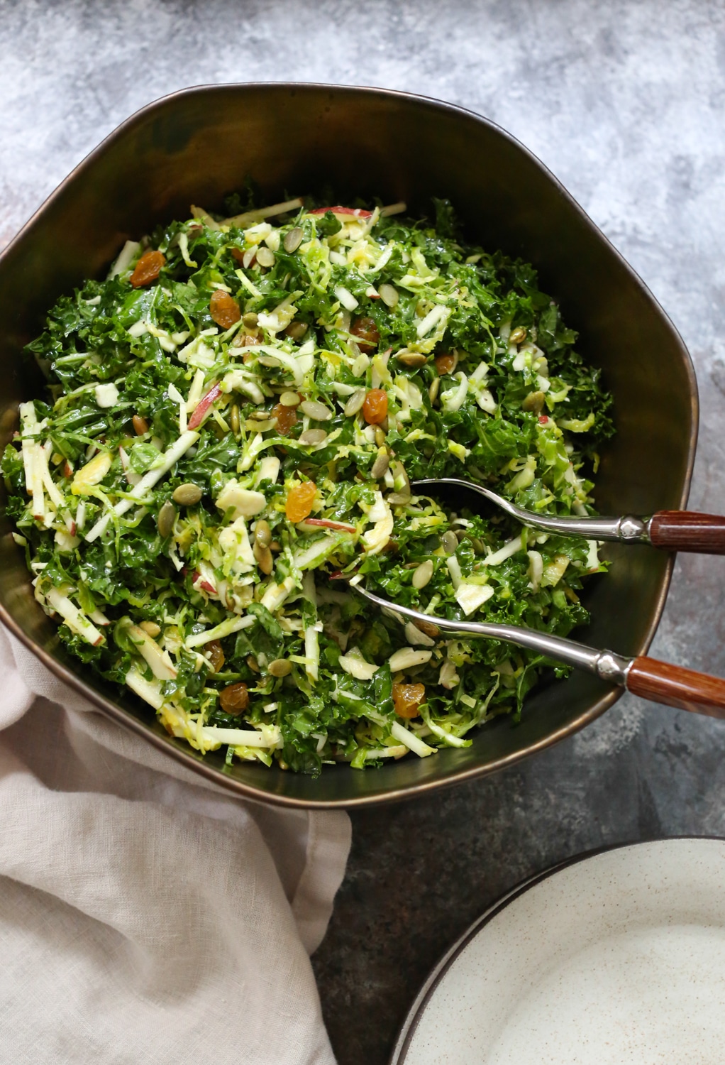 Overhead closeup of Kale, Brussels Sprout, and Apple Salad in a bowl with serving utensils in the bowl and side plates in the bottom right corner.