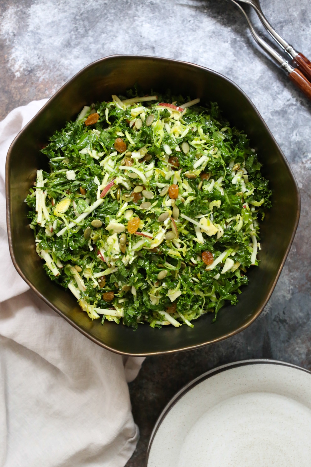 Overhead view of Kale, Apple, and Brussels Sprouts Salad in a large bowl with some serving utensils peeking into the upper right corner.